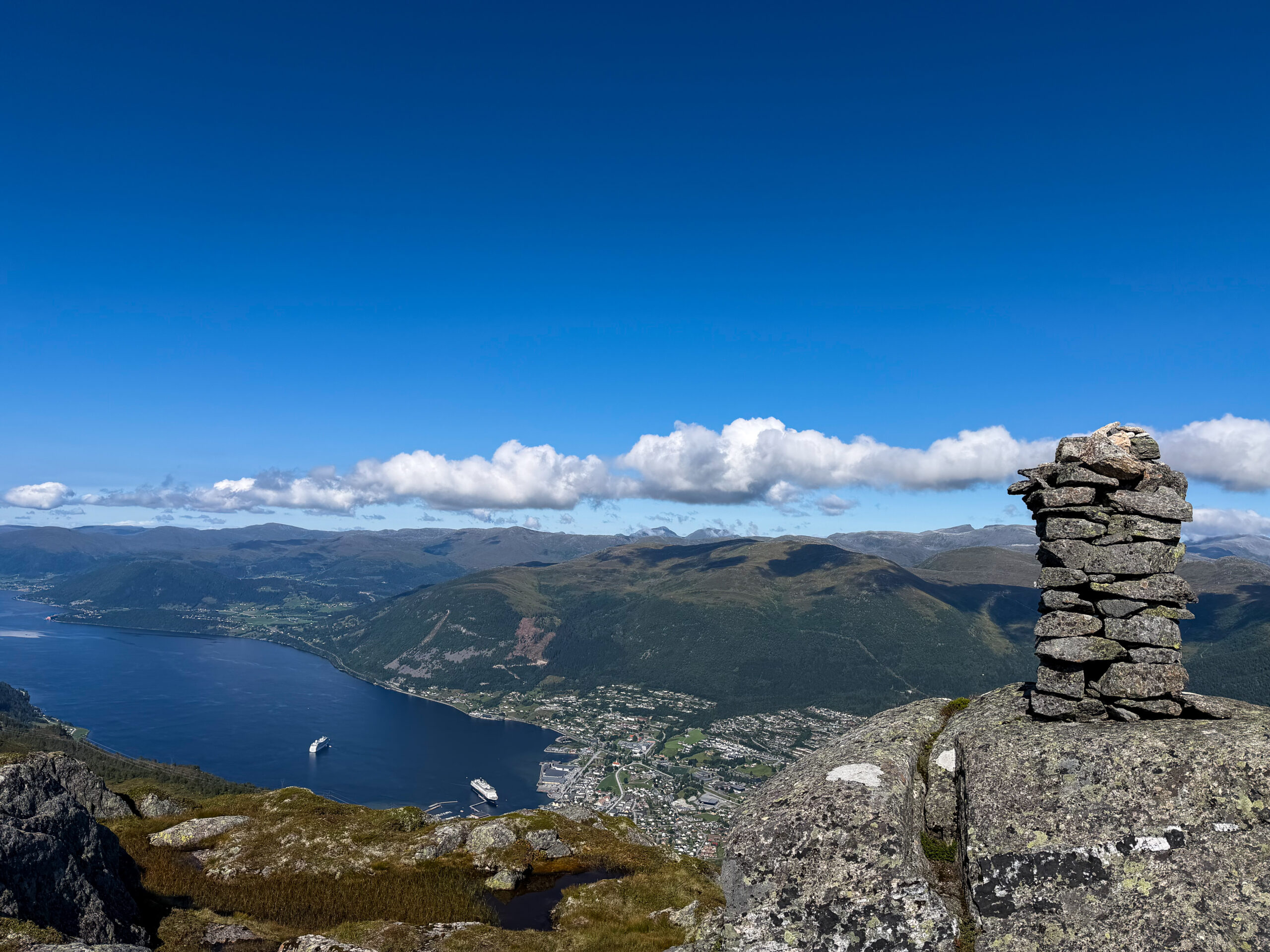 Stone cairn on a summit above the fjord, with the town and mountains below.