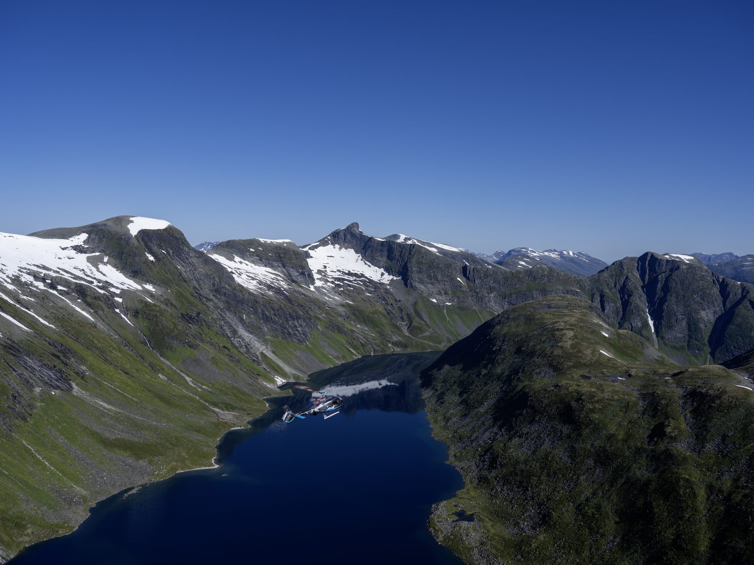 Mountain lake surrounded by snow-streaked peaks, seen from a helicopter.