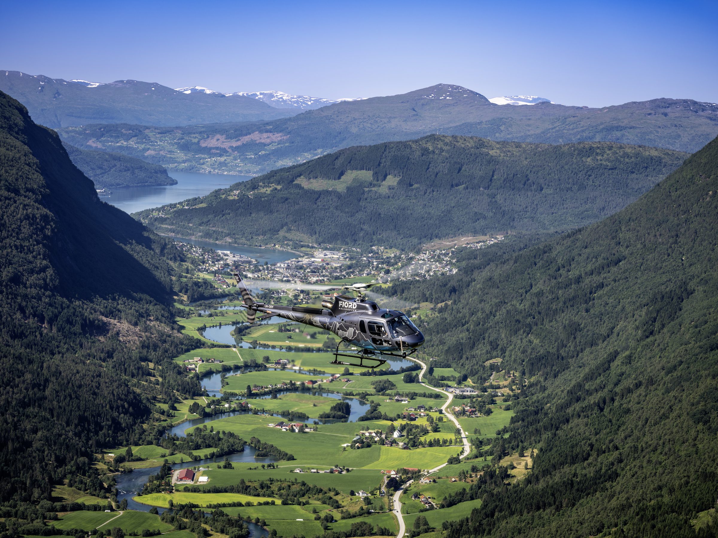 Fjord Helikopter helicopter flying over a green valley and a fjord-side town.